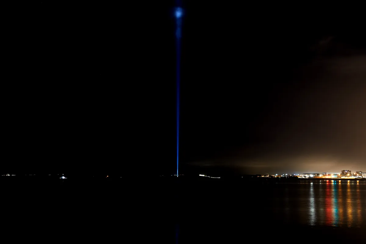 A night view of the Imagine Peace Tower on Viðey Island, Iceland, featuring a powerful beam of blue light extending vertically into the dark sky. The surrounding landscape is shrouded in darkness, with the light from the tower creating a striking contrast. In the distance, the lights of Reykjavik reflect off the calm waters, adding a subtle glow to the horizon. The image captures the serene and contemplative atmosphere of this iconic monument dedicated to peace and harmony.