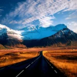 A stunning view of the Ring Road in Iceland, stretching into the distance with snow-capped mountains dominating the horizon. The road is flanked by golden fields, leading the eye towards the rugged and majestic peaks. The sky above is a brilliant blue, scattered with wispy clouds, creating a picturesque and serene landscape that captures the wild beauty of Iceland's natural scenery.