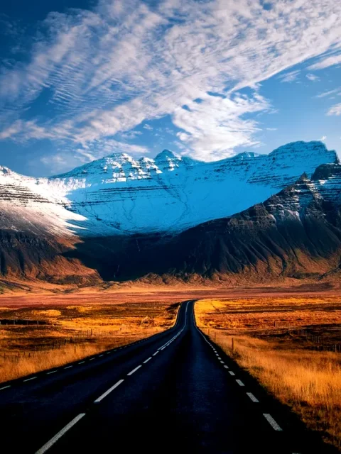 The Ultimate Guide to Iceland’s Ring Road 1 A stunning view of the Ring Road in Iceland, stretching into the distance with snow-capped mountains dominating the horizon. The road is flanked by golden fields, leading the eye towards the rugged and majestic peaks. The sky above is a brilliant blue, scattered with wispy clouds, creating a picturesque and serene landscape that captures the wild beauty of Iceland's natural scenery.