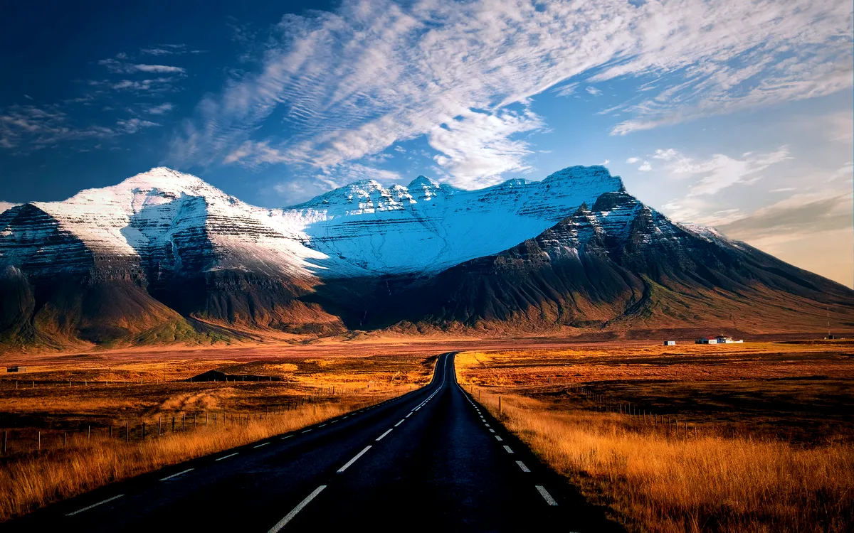 The Ultimate Guide to Iceland’s Ring Road 1 A stunning view of the Ring Road in Iceland, stretching into the distance with snow-capped mountains dominating the horizon. The road is flanked by golden fields, leading the eye towards the rugged and majestic peaks. The sky above is a brilliant blue, scattered with wispy clouds, creating a picturesque and serene landscape that captures the wild beauty of Iceland's natural scenery.
